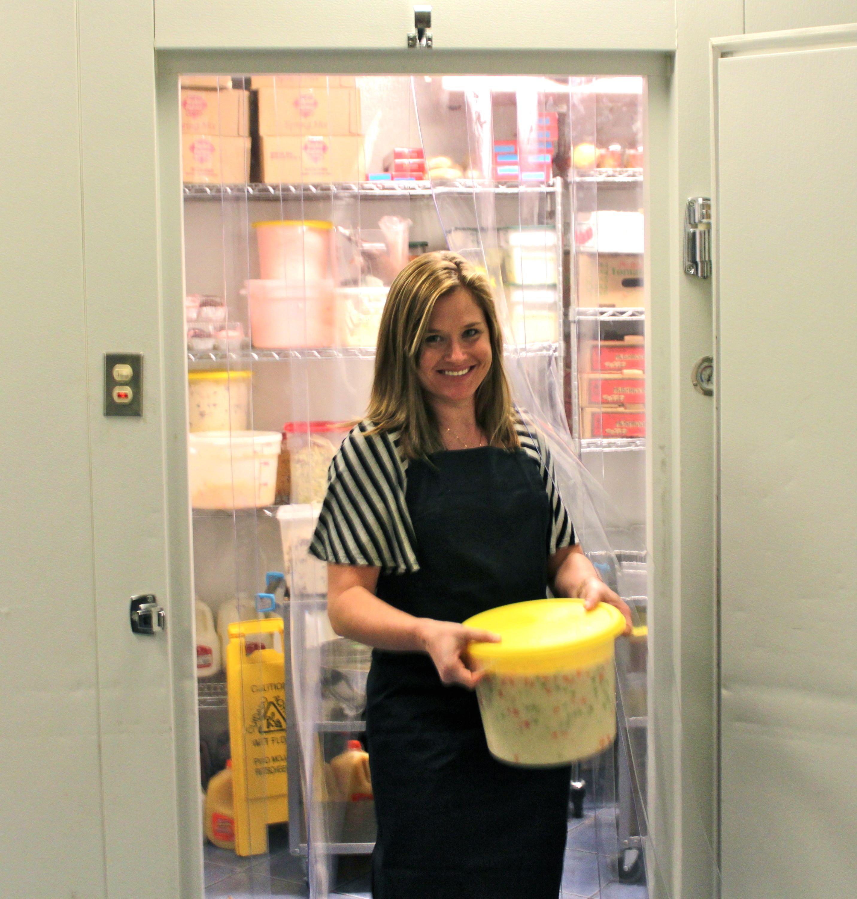 woman holding a food container while exiting a walk-in refrigerator through clear PVC strip curtains in a commercial kitchen