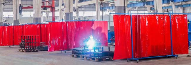 Welder working behind red Screenflex PVC curtains in an industrial facility with multiple mobile partitions