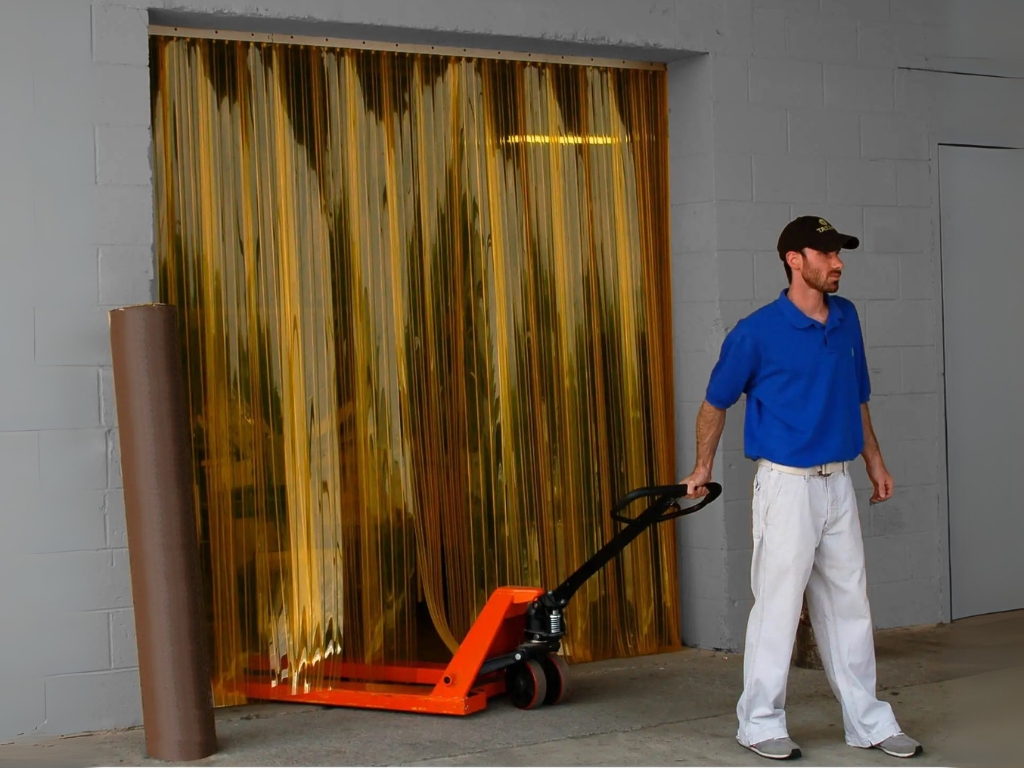 A man with a pallet jack stands next to a yellow PVC strip curtain.