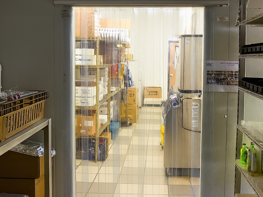 The interior of a walk-in cooler seen from the outside. Clear plastic strip curtains hang in the doorway. Shelves are visible inside, filled with various boxes and containers. A refrigerator is on the right side.