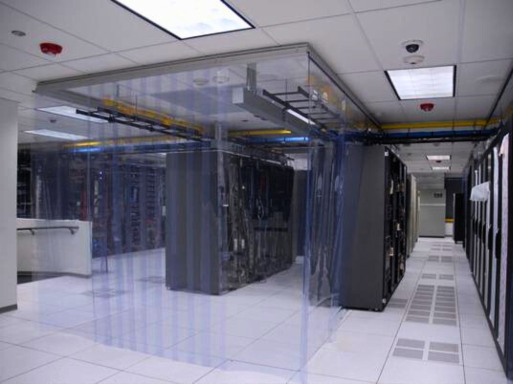 Inside a data center, a large cube-shaped enclosure made of clear plastic strips surrounds a section of server racks.