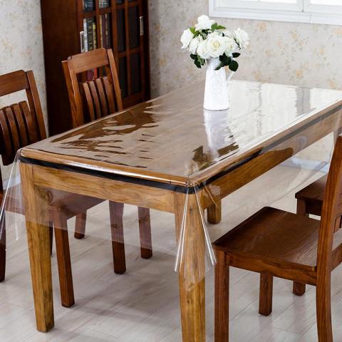 Wooden dining table with four chairs covered by a clear protective vinyl sheet, decorated with a vase of white flowers.