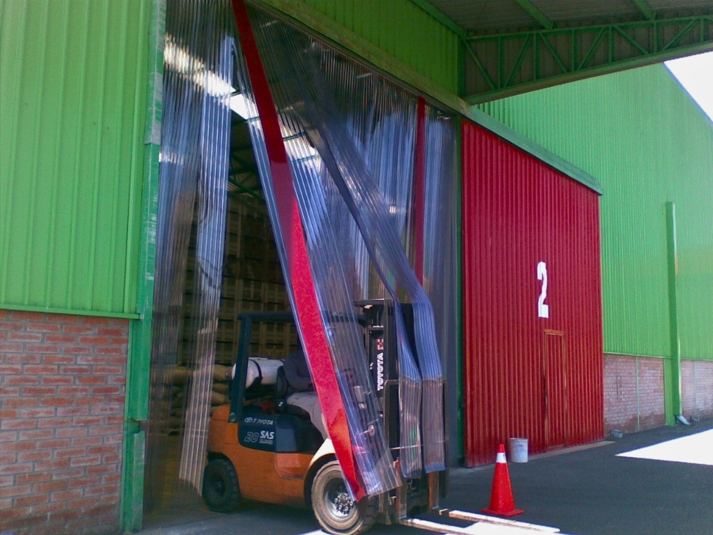 An orange forklift drives through a doorway with fire-retardant clear plastic strip curtains. The building has a green corrugated metal upper wall and a red corrugated lower wall with the number '2' painted on it. A single orange traffic cone is on the ground to the right of the forklift.