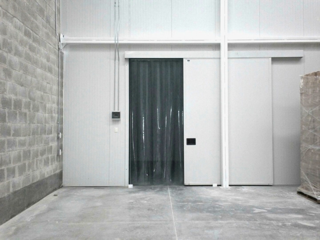 A wide shot of a warehouse with a large gray brick wall on the left. In the center are two white cold storage doors, one of which is a sliding door with clear plastic strip curtains hanging inside the opening.