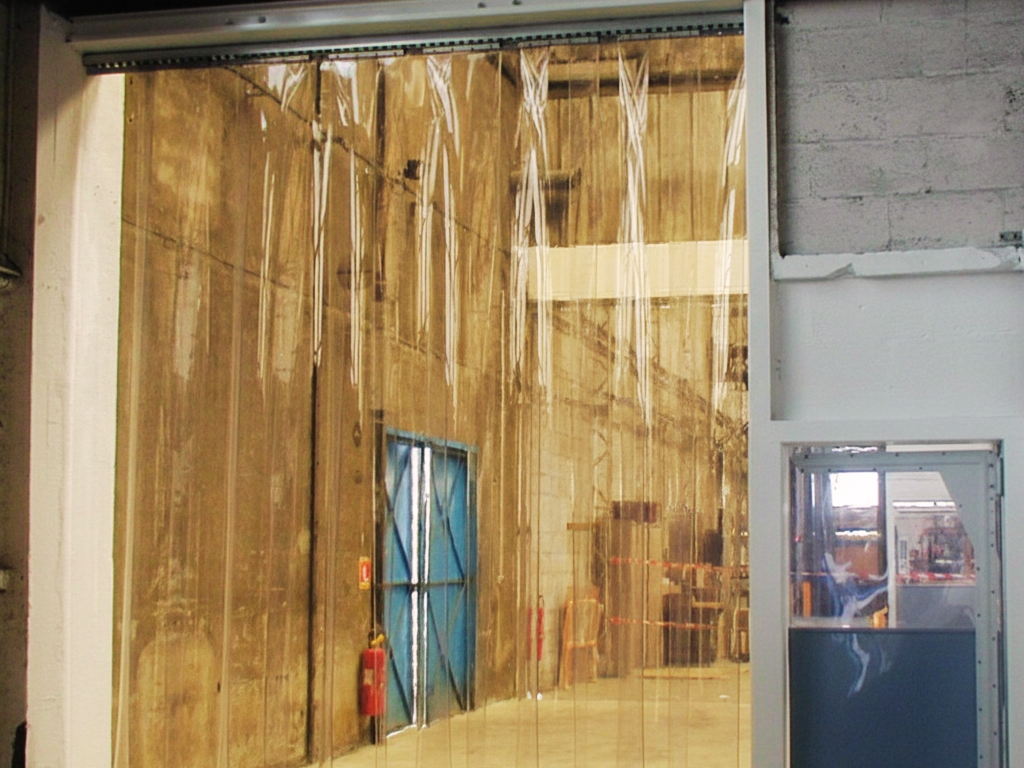 A large opening in a warehouse is covered by clear plastic strip curtains. Through the curtains, another large, industrial space with concrete walls and a blue metal door can be seen.