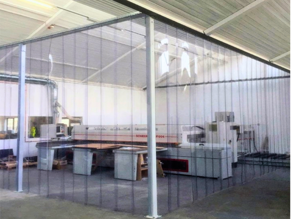 A large piece of industrial machinery is enclosed within a clear PVC strip curtain barrier, which is suspended from the ceiling of a warehouse or workshop.
