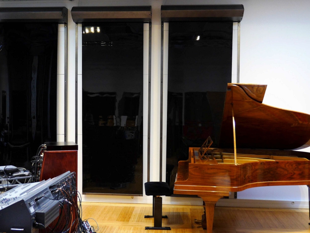 A grand piano placed in a music studio with tall black acoustic panels and sound equipment nearby.