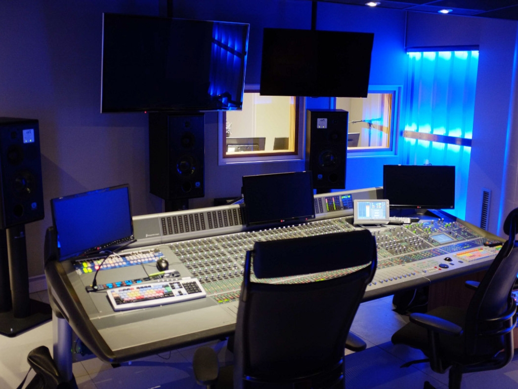 Modern recording studio control room with large mixing console, multiple monitors, and ambient blue lighting.