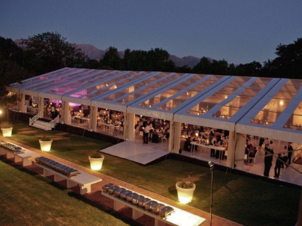 A large, modern-looking clear-topped tent is set up for an outdoor wedding reception. Tables and guests are visible inside the lit-up structure at night. Buffets with chafing dishes are set up outside on the grassy lawn.