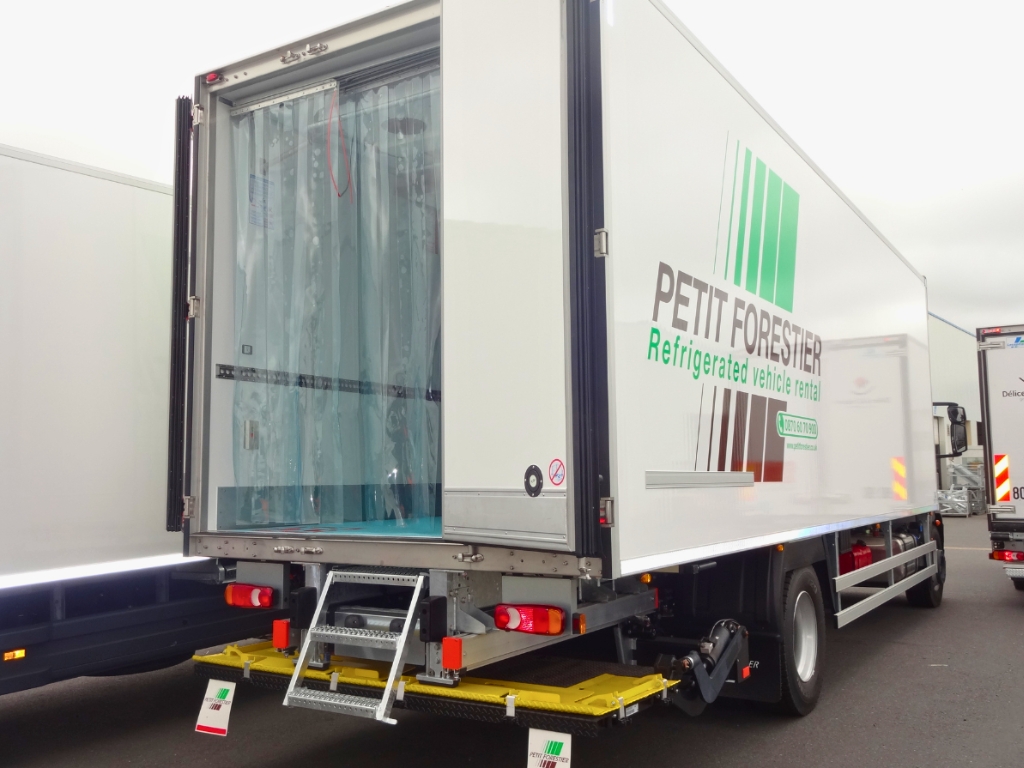 The back of a "Petit Forestier" refrigerated rental truck. The door is open, revealing clear plastic strip curtains hanging inside the opening. A metal ladder is folded down from the truck bed.
