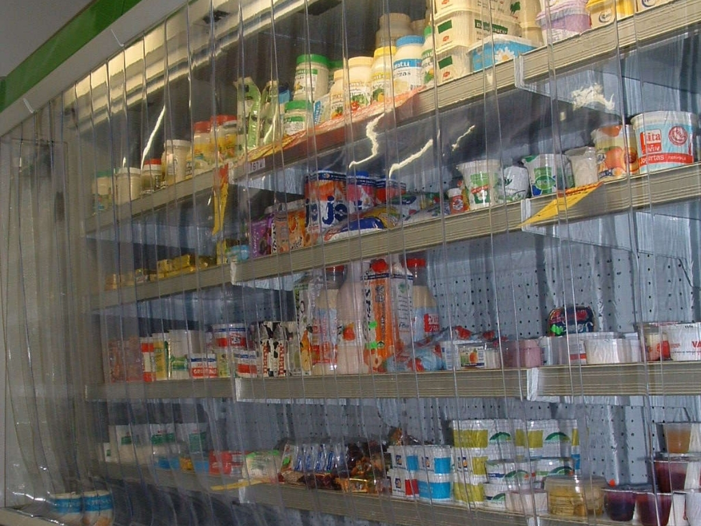 A clear plastic strip curtain hangs in front of a supermarket chiller aisle, covering shelves stocked with various dairy products and other refrigerated goods.