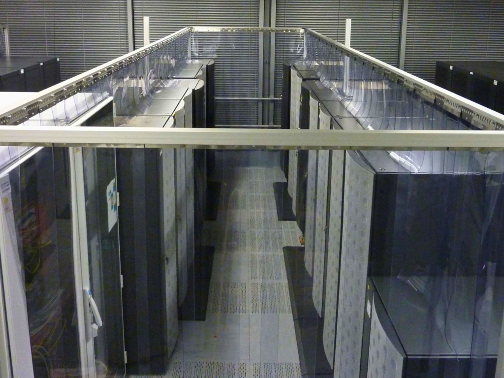 An overhead view of a data center aisle with two rows of tall, dark server racks. The aisle is enclosed by clear plastic panels and a metal frame, creating a hot or cold aisle containment system.