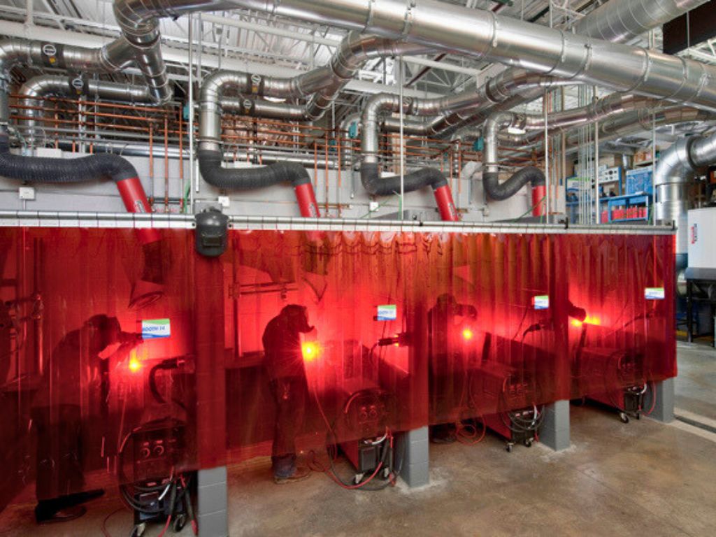 Row of welders working behind red strip curtains with ventilation pipes overhead.