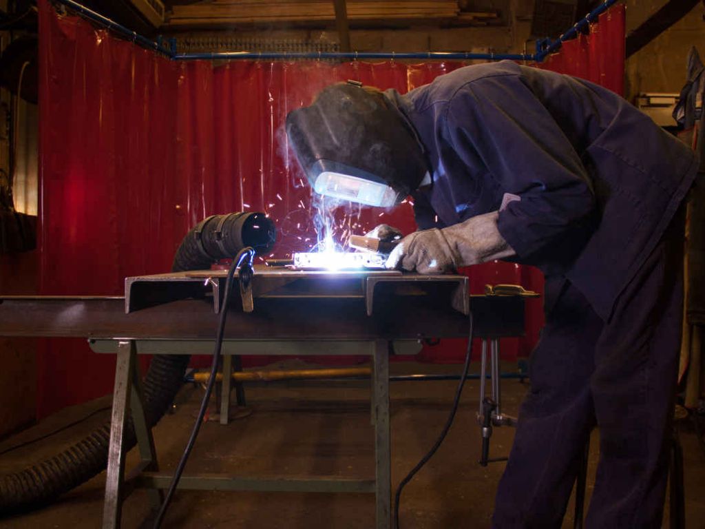 Worker welding metal with protective helmet and gloves behind red safety screen.