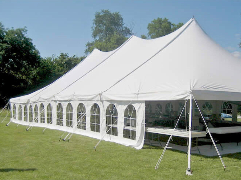 A long, white pole tent with clear arched windows along its side wall, set up on a grassy lawn. The sky is clear, and a line of trees is in the background.