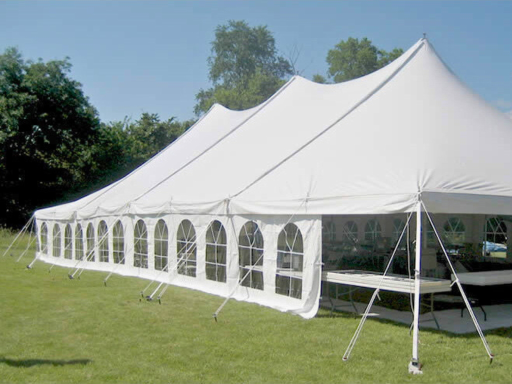A long, white pole tent with clear arched windows along its side wall, set up on a grassy lawn. The sky is clear, and a line of trees is in the background.