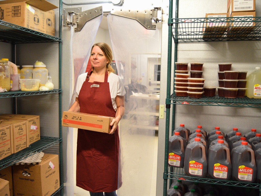 Woman in apron carrying a box through transparent swing pvc doors in a storage room.