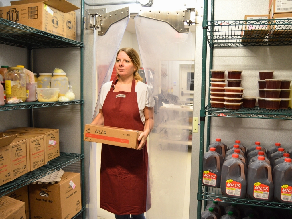 A woman wearing a maroon apron stands in the doorway of a walk-in cooler, holding a cardboard box. Clear plastic strip curtains hang behind her. The interior has wire shelving filled with food and beverages.