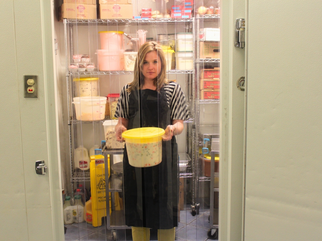 A woman in an apron stands inside a walk-in cooler, holding a large yellow lidded tub of food. Clear plastic strip curtains hang in the doorway in front of her.