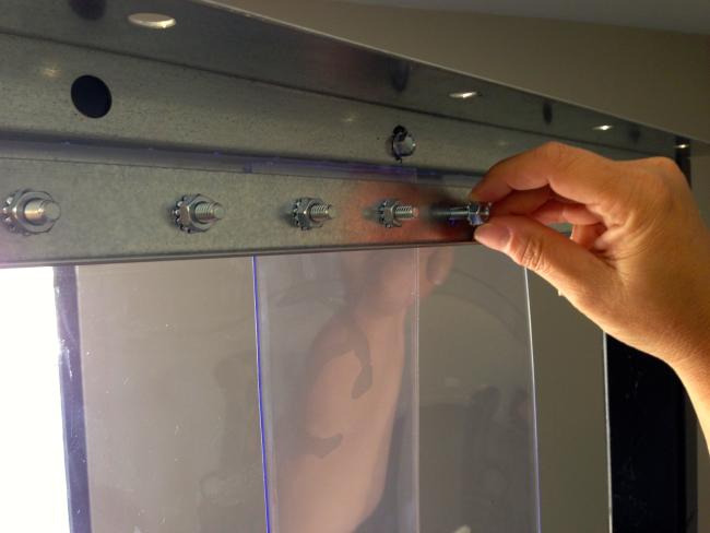 A detailed shot of a hand tightening a bolt on a metal mounting bracket, securing a clear vinyl curtain in place with a sturdy galvanized steel structure.