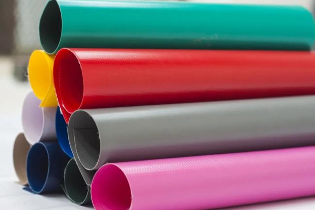 A close-up view of stacked rolls of PVC tarpaulin in various vibrant colors, including red, blue, green, and gray, with a smooth, flexible texture.